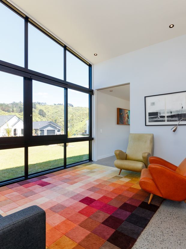Bright contemporary living room with a dramatic floor-to-ceiling black-framed window overlooking a green hillside and neighbouring homes. The room has white walls and ceiling, polished concrete flooring, a large multicoloured patchwork rug, a mustard lounge chair, an orange leather armchair, a dark sofa edge, framed artwork, and a slim floor lamp. Recessed ceiling lights and the expansive glazing create an airy, colourful interior.