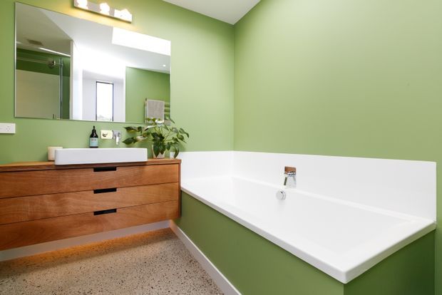 A contemporary bathroom with a vivid sage-green painted wall, a white built-in bathtub, and a warm timber floating vanity with a white vessel basin. A large frameless mirror reflects the shower enclosure and window, while terrazzo-style flooring and under-vanity lighting add texture and warmth.
