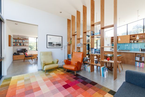 Bright open-plan living and dining space with a colourful patchwork rug, two leather lounge chairs, extensive timber open shelving and room-dividing posts, polished concrete flooring, and large windows. A compact kitchen with timber cabinetry and a blue glass splashback is visible in the background, while a separate sitting or media area opens to the left.
