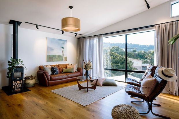 Warm, contemporary living room with extensive floor-to-ceiling glazing overlooking a leafy suburban landscape. The room features a leather sofa and matching lounge chair, sculptural timber coffee table, area rug, indoor plants, a black freestanding wood-burning stove, track lighting, and a large woven pendant light. Timber flooring and neutral furnishings create a relaxed, inviting atmosphere.
