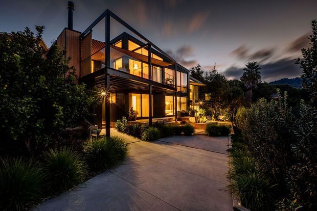 Twilight exterior view of a contemporary multi-level home set within dense landscaped planting. The house features dark structural framing, extensive floor-to-ceiling glazing, warm timber cladding and soffits, projecting balconies, and a prominent steep gable-like roof form. A broad concrete path leads toward the illuminated entrance and outdoor living areas.