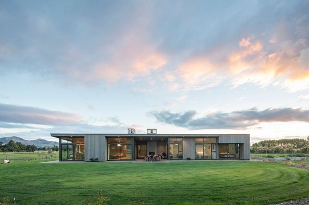 A low, single-storey contemporary residence set in a broad landscaped lawn, with a long rectangular form, extensive floor-to-ceiling glazing, dark window frames, and vertically ribbed grey metal cladding. The house opens toward the lawn with a sheltered central outdoor seating area. Rolling hills, cultivated fields, and a dramatic sunset sky form the distant background.