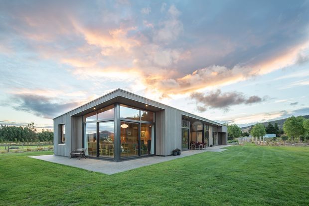 A low, single-storey contemporary house set in a spacious landscaped lawn, photographed from the exterior at dusk. The building has a simple rectangular form, broad flat roof overhangs, extensive floor-to-ceiling glazing, dark metal-framed windows and doors, and grey vertical exterior cladding. A sheltered outdoor seating area is visible along the side, with rural vegetation and hills in the background.