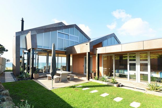 Contemporary coastal residence with angular pitched roof forms, extensive floor-to-ceiling glazing, dark metal cladding and warm timber-toned wall panels. The image is viewed from a landscaped lawn and paved outdoor dining terrace, with the ocean visible beyond the house.