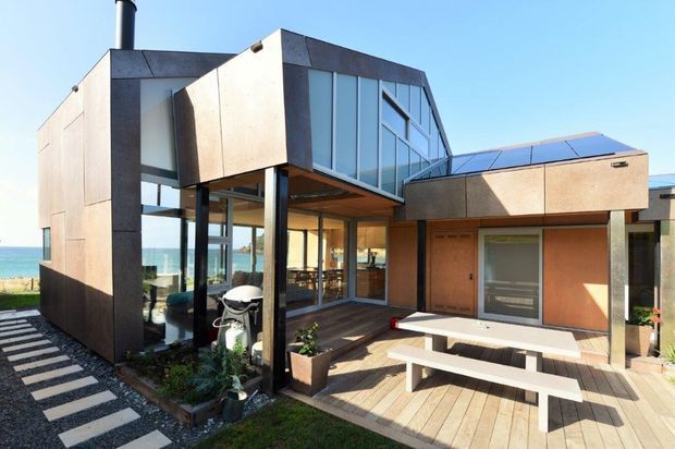 Contemporary multi-level coastal home with strong geometric forms, dark metal cladding, expansive glazing, and a timber-decked courtyard. The elevated central volume features angled glazing and a projecting canopy, while the lower wing includes a garage or utility door. A paved stepping-stone path, built-in bench, outdoor dining table, and planting surround the home, with the ocean visible beyond.