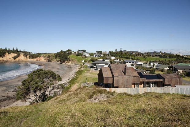 Wide elevated exterior view of a coastal residential property beside a sandy beach and shoreline. The prominent house has dark timber-clad pitched roof forms, large glazed openings, and a pale boundary fence, with neighbouring homes and rolling green landscape in the background under a clear blue sky.