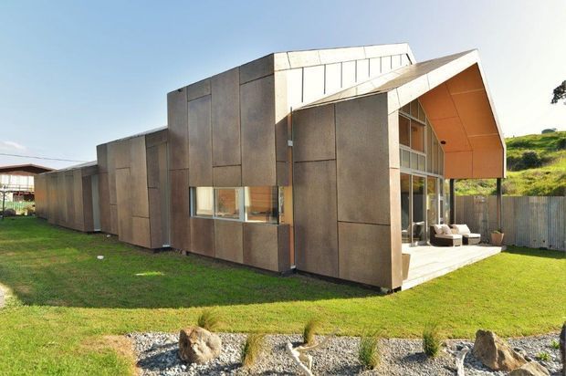 Exterior view of a contemporary single-storey house with angular, folded roof forms and dark vertical-panel cladding. Large glazed openings connect the living area to a small paved outdoor space, while the home sits within a grassy landscaped site.