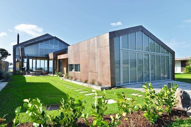 Exterior view of a contemporary, single-storey house with a prominent glazed gabled volume, rust-toned metal or weathered steel wall cladding, dark structural framing, and extensive floor-to-ceiling windows. The building sits in a landscaped suburban garden with lawn, planting beds, and stepping-stone paths under bright daylight.
