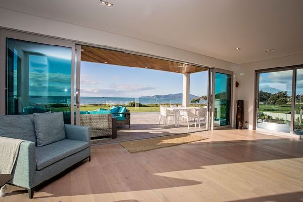 Bright contemporary living room with pale timber flooring, a blue-grey armchair, and expansive sliding glass doors opening onto a covered outdoor terrace. The terrace includes outdoor seating and dining, with views across a pool or lawn toward a coastal landscape and hills.
