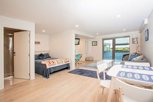 Bright open-plan coastal-style living and dining area with pale timber flooring, white walls, recessed ceiling lights, and large windows opening toward a water view. The space includes a dining table with upholstered white chairs, a compact white coffee table, a grey sofa, a blue-toned rug, and a bed or daybed positioned near the left side. A bathroom or utility room is visible through the open door.