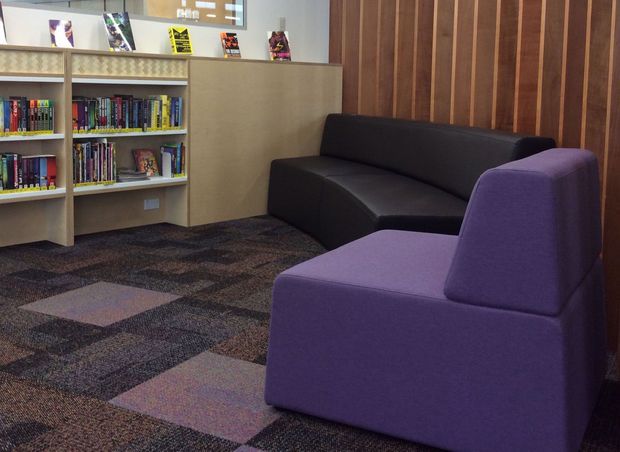 A compact contemporary reading lounge or library with open built-in bookshelves, a pale wood display cabinet, dark and purple upholstered seating, and a patterned carpet. Vertical timber wall panelling forms a warm feature wall behind the sofas, while books and magazines occupy the shelving.