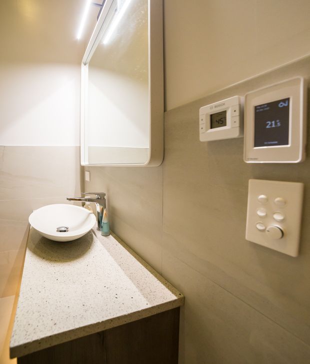 Compact contemporary bathroom vanity with a white vessel basin, chrome tapware, speckled light benchtop, dark wood-toned cabinetry, mirrored wall cabinet, and beige tiled wall finish. Digital controls and a wall-mounted display are visible beside the vanity.