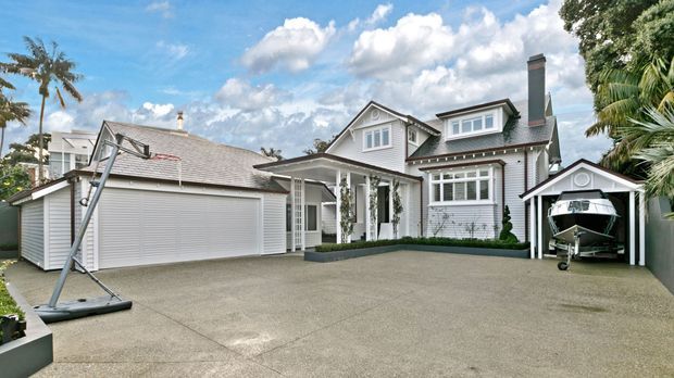 Exterior view of a substantial light-grey and white multi-storey home with a pitched gabled roof, dormer windows, covered entry porch with columns, attached garage, and a separate boat storage structure. The broad concrete driveway dominates the foreground, with mature tropical landscaping around the property.