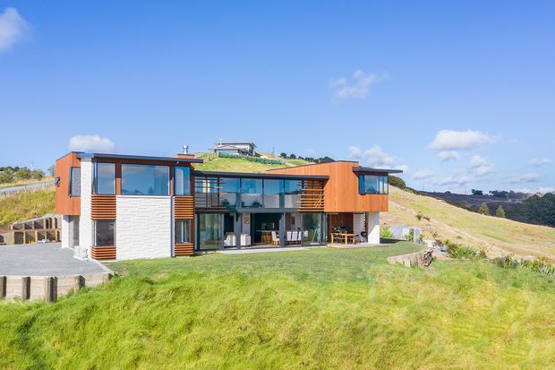 A contemporary multi-level house set on a sloping grassy site, with extensive glazing, projecting orange-brown timber-clad wings, horizontal timber sun-shading screens, and a central glazed living area. The home has a strong horizontal composition and views across an open rural or coastal landscape.