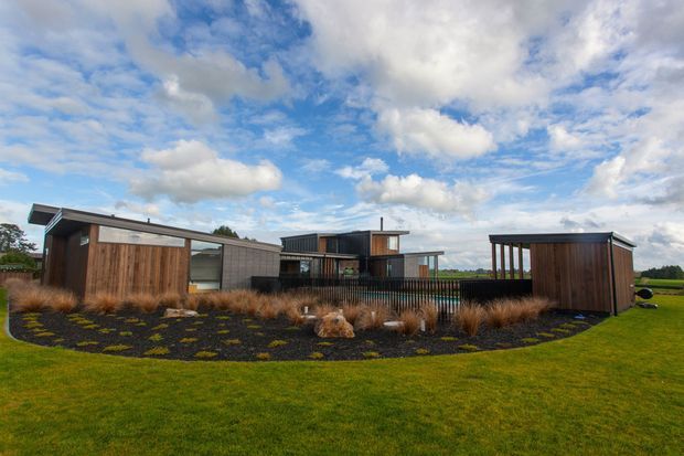 Low, contemporary residential buildings arranged around a landscaped outdoor area, featuring dark timber cladding, concrete block walls, broad roof overhangs, and extensive glazing. Ornamental grasses and dark mulch surround the structures, with a manicured lawn and open rural-looking landscape beyond under a partly cloudy sky.