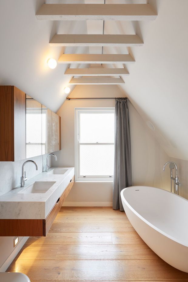 Bright attic-style bathroom with a freestanding white bathtub, double vanity, wall-mounted mirrors, and brushed metal tapware. The room has a pitched sloping ceiling with exposed white-painted beams, a central window with grey curtains, warm timber flooring, and wood-grain vanity cabinetry with a pale stone benchtop.