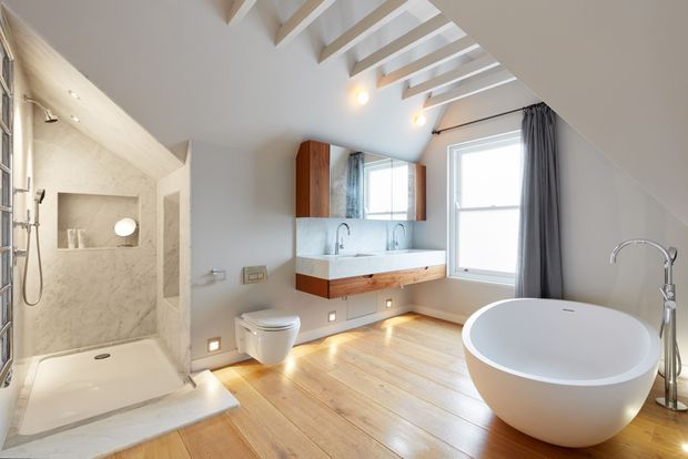 Bright, spacious contemporary bathroom with a freestanding white oval bathtub, walk-in shower, wall-hung toilet, double vanity with timber cabinetry, pale stone or marble shower surfaces, timber flooring, and a sloped ceiling with exposed white beams. Large windows and skylights provide abundant natural light.