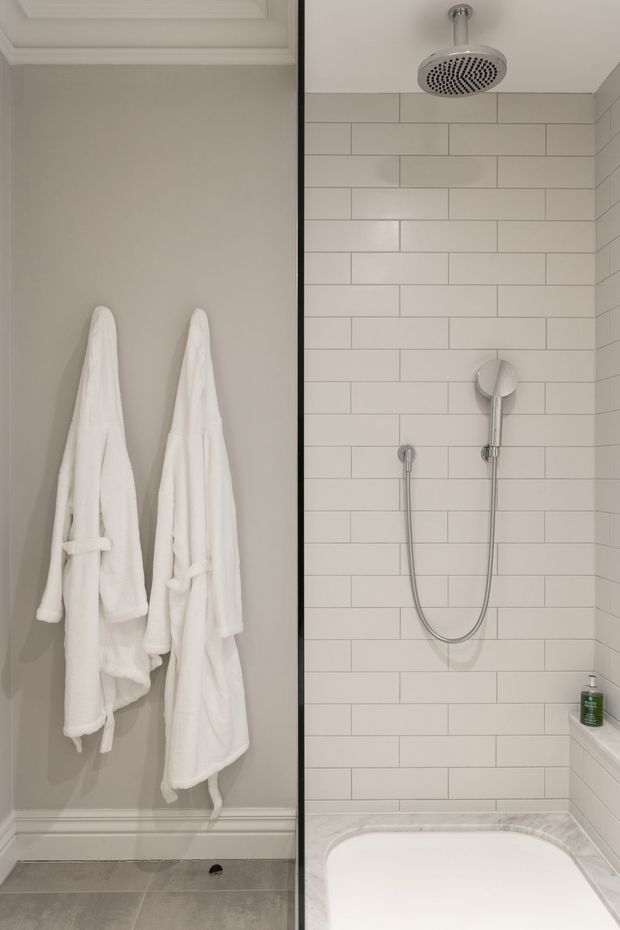 A compact, refined bathroom featuring a walk-in shower and bathtub combination. The shower walls are clad in light rectangular subway tiles with contrasting grout, complemented by chrome fixtures and a white tub. On the left, two white bathrobes hang against a warm light-grey painted wall with decorative trim and moulding. The overall palette is neutral and softly lit.