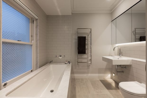 Bright contemporary bathroom with a white built-in bathtub beneath frosted patterned glazing, white subway-tile walls, a floating white vanity with integrated basin, mirrored cabinetry, chrome tapware, heated towel rail and toilet. Light timber-look flooring and concealed LED lighting create a clean, refined feel.