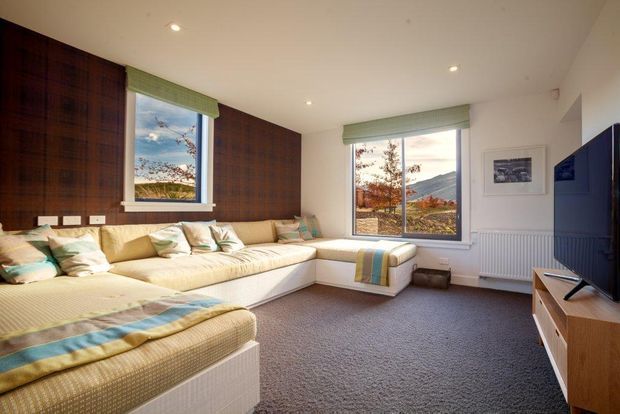 A bright, cozy living room with a large L-shaped upholstered built-in window seat or sectional, layered cushions and throws, dark timber wall panelling, pale walls, grey carpet, and a timber TV console. Two windows with green Roman blinds overlook a mountainous landscape, while recessed ceiling lights provide even illumination.