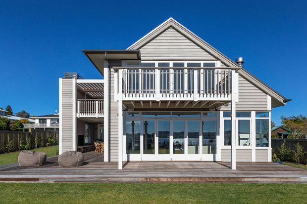 Exterior rear view of a light grey weatherboard house with a prominent gabled roof, upper-level balcony, extensive white-framed glazing, and a broad timber deck overlooking a lawn. The design has a coastal, relaxed character with strong indoor–outdoor connection.