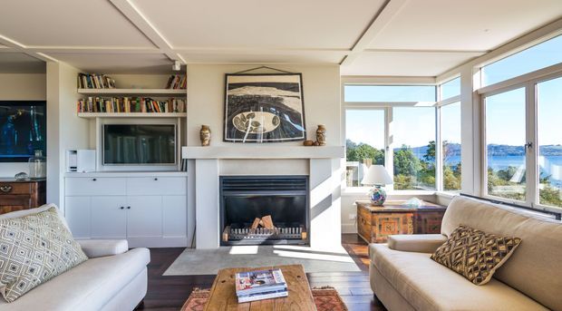 Bright, refined living room with a central fireplace and mantel, built-in white cabinetry and open bookshelves, a television, upholstered sofas, a rustic timber coffee table, and extensive corner glazing overlooking a blue-water landscape. The ceiling has clean recessed panel detailing and the room receives abundant natural light.