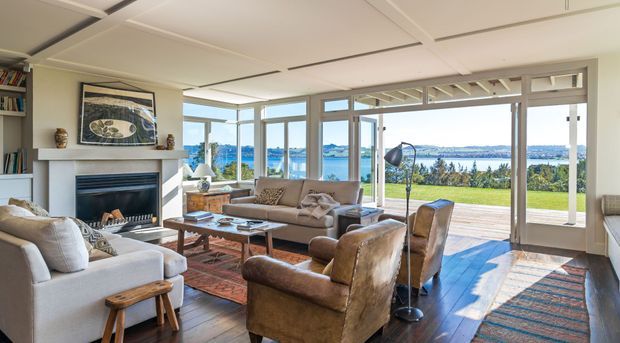 Bright, spacious living room with a fireplace, sofas and leather armchairs arranged around a coffee table. Large floor-to-ceiling glazed doors and windows open the room to a timber deck and expansive waterfront view. The room has dark timber flooring, a light fireplace surround, a patterned area rug, and a white suspended-panel ceiling.