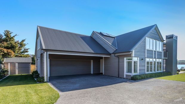 Exterior view of a contemporary multi-gabled house with grey weatherboard cladding, corrugated metal roofing, large white-framed windows, and an integrated double garage. A separate small shed, landscaped lawn, driveway, and a glimpse of water are visible.
