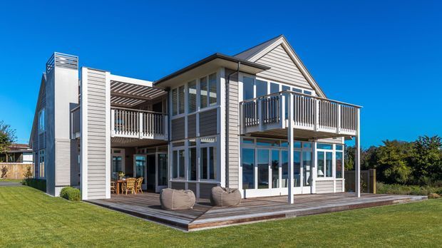 A two-storey coastal-style home viewed from the rear, featuring light weatherboard cladding, a pitched gable roof, extensive white-framed glazing, upper and lower verandah areas, a sheltered outdoor dining space, and a broad timber deck overlooking a lawn.