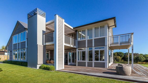 Contemporary multi-level residential house with a strong vertical composition, grey and white weatherboard cladding, extensive glazing, balconies, a timber deck, and a prominent tall chimney or service tower. The home opens onto a neatly landscaped lawn and appears to sit on a gently elevated site with distant trees beyond.