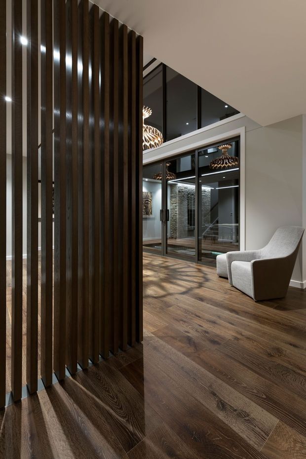 A sophisticated contemporary interior hallway or open living area with dark timber flooring, a dramatic vertical timber slat divider, expansive black-framed glazing, and sculptural pendant lights. A light upholstered lounge chair sits beside the glazed partition, while reflections reveal additional seating and textured feature walls beyond. The space has a refined, moody atmosphere with strong linear architectural detailing.