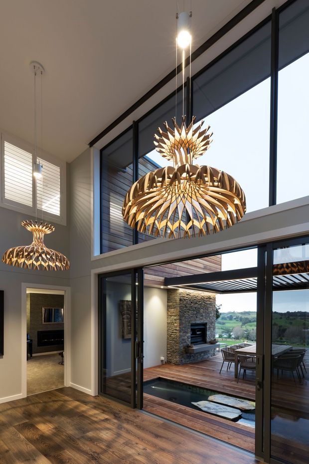 Double-height contemporary interior opening onto an outdoor deck and landscape, with extensive black-framed glazing, dark timber flooring, and distinctive sculptural wooden pendant lights. A stone outdoor fireplace, dining area, water feature, and rolling green landscape are visible beyond the open doors.