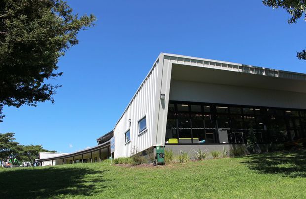 Low-slung modern building viewed from a gently sloping grassy site under a clear blue sky. The structure has a long horizontal form, a broad projecting flat roof, extensive dark-framed glazing, pale vertical metal cladding, and a covered outdoor edge with landscaped planting. Trees frame the scene.