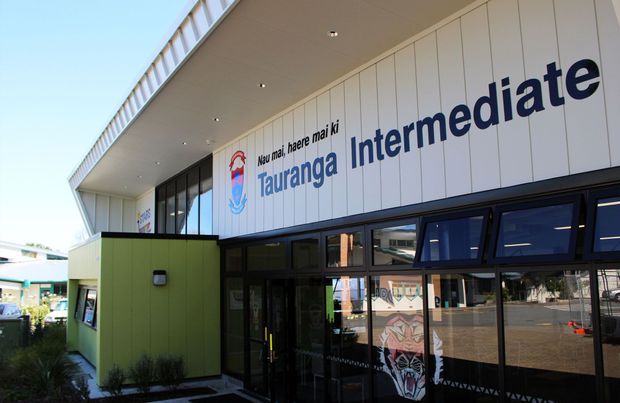 Exterior view of the Tauranga Intermediate school entrance, featuring a contemporary white-panelled facade with large bilingual signage, dark glazed entry doors and windows, a projecting flat canopy, and a green entrance volume.