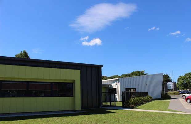 Exterior view of a low-rise contemporary commercial or institutional building complex. The foreground structure has a dark metal-clad rectangular volume with a vivid olive-green panelled facade and long horizontal glazing, while a lighter corrugated metal-clad wing extends behind it. A paved pedestrian path, lawn, landscaping, and parked cars are visible under a bright blue sky.
