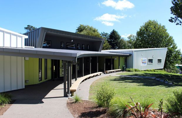 Exterior view of a low-rise contemporary institutional or commercial building arranged around a landscaped courtyard. The complex features corrugated metal cladding, dark glazed upper-level volumes, a broad flat or gently sloping roof, covered walkways, and a curved timber bench along the paved path. Bright green and pale grey facade panels contrast with dark charcoal elements, with mature trees and planting around the site.