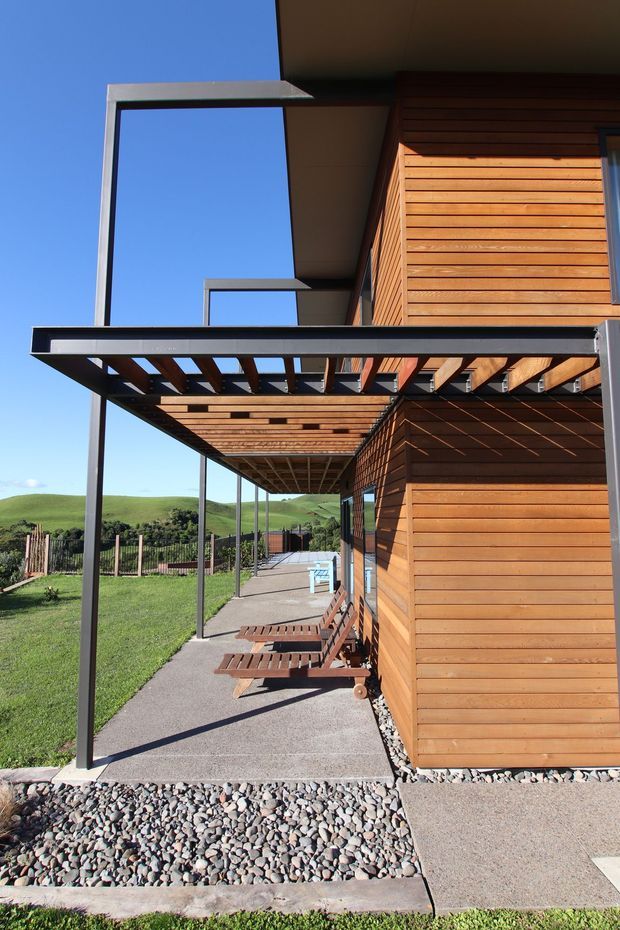 Exterior architectural view of a contemporary building with warm horizontal timber cladding, dark metal framing, and a deep pergola/verandah formed by timber slats and steel posts. A concrete walkway runs alongside the building, with gravel drainage landscaping and rolling green hills visible beyond.