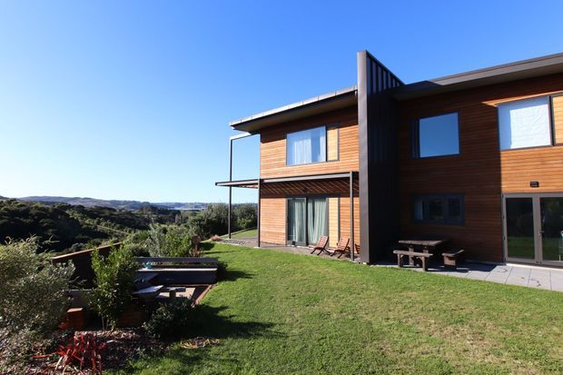 Exterior view of a contemporary two-storey timber-clad house set on a spacious grassy site, with large windows, a projecting upper-level balcony or deck, paved outdoor seating area, and expansive rolling landscape views. The building has a strong vertical dark feature element separating the main timber volumes and a simple low-pitched roofline.
