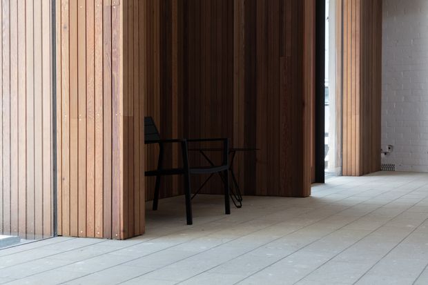 Interior view of a refined contemporary space with extensive vertical timber wall panelling and matching timber screens or portals. A small black dining or work table and chairs sit against the dark timber backdrop. The room has a pale tiled floor, a white-painted brick wall, and strong natural light entering through a narrow opening, creating a warm yet restrained architectural atmosphere.
