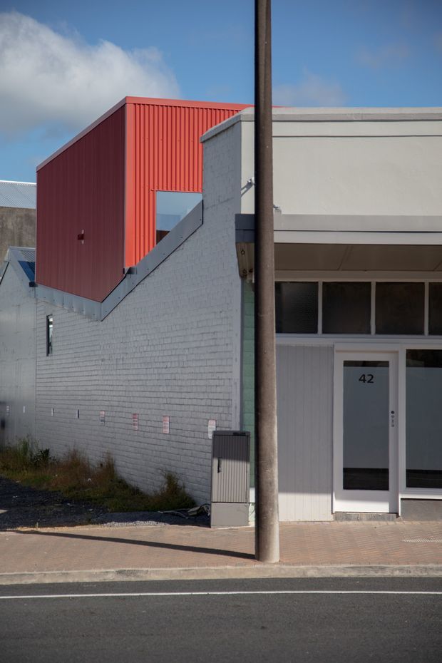 Street-facing view of a compact urban commercial or mixed-use building. The facade combines a white-painted brick side wall with a projecting red corrugated-metal upper volume and a simple white entrance frontage marked with the number 42. A paved footpath and roadway occupy the foreground, while neighboring low-rise structures are partially visible.