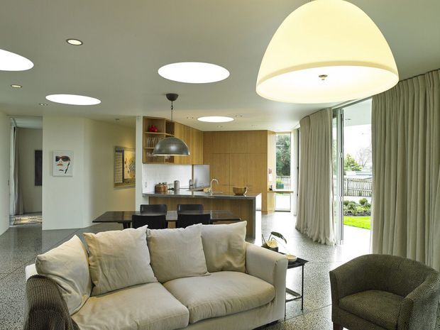 Open-plan contemporary living area with a large cream sectional sofa in the foreground, a textured armchair, and a compact kitchen with timber cabinetry and island behind it. Full-height glazed doors with sheer curtains open to a green outdoor area. The space has a polished terrazzo floor, white ceiling with multiple recessed and pendant lights, and a bright, relaxed atmosphere.