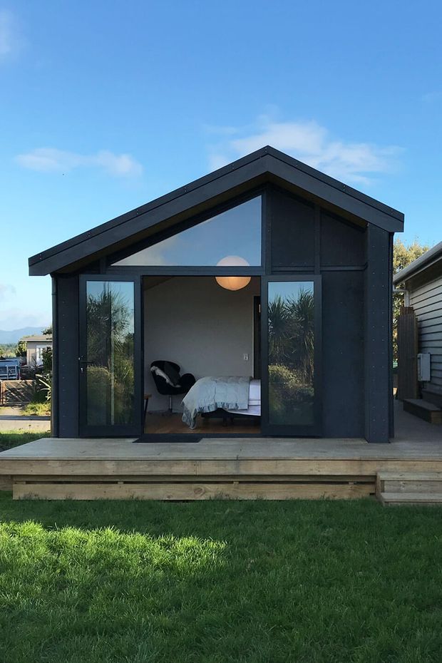 Exterior view of a compact dark-clad detached backyard studio or small dwelling on a raised timber deck. The simple gable-front building has large glazed sliding doors, a triangular clerestory window, dark vertical cladding, and a visible bedroom interior with a bed, lounge chair, and pendant light. Grass and neighbouring structures surround the deck.