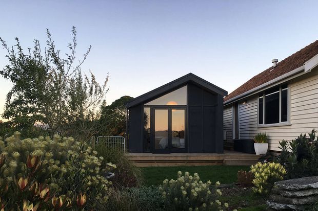 Exterior view of a compact dark-clad garden studio or extension beside a weatherboard house. The studio has a simple pitched roof, large glazed entry doors, and a timber deck, set within a landscaped garden with native-style planting. The adjacent house has light weatherboard cladding, white-framed windows, and a tiled pitched roof.