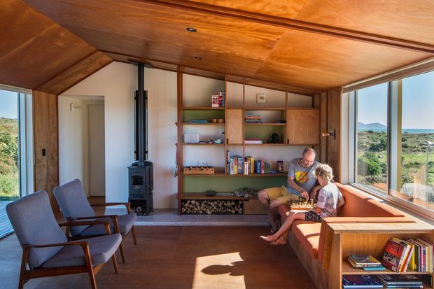 Warm, compact living room with extensive timber lining across the ceiling and built-in shelving wall. The room includes a wood-burning stove, upholstered lounge chairs, a built-in window seat or sofa, and large glazed windows with views to the surrounding landscape and water. The space has a mid-century-influenced, cabin-like character with integrated timber joinery and abundant natural light.