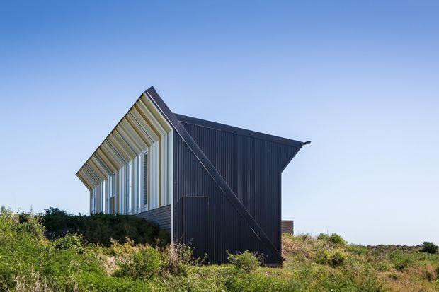 Exterior view of a sharply angular, contemporary single-storey building set within low coastal-style vegetation. The structure has dark corrugated metal cladding, a pronounced pitched or folded roof form, and a glazed gable facade with projecting vertical fins or framing elements. Clear blue sky and open scrub landscape surround the building.