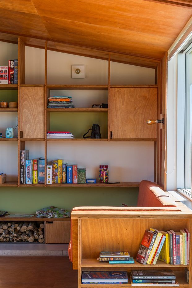 A warm, compact living room or reading nook with extensive custom timber shelving and cabinetry, filled with books, board games, and small objects. The built-in joinery continues across the wall and around a low divider, with a large window bringing in strong natural light. A green painted wall, timber ceiling, wood flooring, and a partially visible rust-coloured upholstered chair create a cosy, mid-century-inspired atmosphere. A built-in log storage niche is visible beneath the shelving.