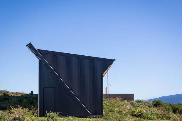 A compact dark-clad building viewed externally against a clear blue sky and low coastal or rural vegetation. The structure has a sharply asymmetrical sloping roofline, corrugated vertical wall cladding, a simple dark door, and a prominent diagonal structural or roof-edge element across the facade. A small timber-toned element is visible beneath the roof edge, with distant hills in the background. The image has a crisp, minimalist contemporary architectural character.