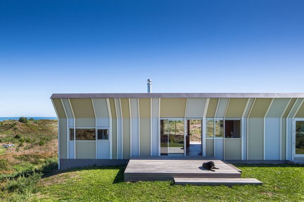A single-storey coastal house viewed from the rear, with a long low facade featuring rhythmic vertical and angled wall cladding panels, extensive glazing, and a small timber deck opening onto a grassy landscape. The ocean is visible in the distance beyond low coastal vegetation under a clear blue sky.