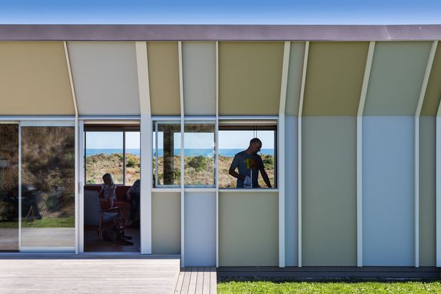 Exterior view of a contemporary single-storey building with a long glazed facade, sliding glass doors, and vertical projecting fins dividing pale green and light grey wall panels. A timber deck and lawn sit in front, while the glazing reveals a small interior seating area and a person. Coastal vegetation and blue water or sky are visible beyond the building.