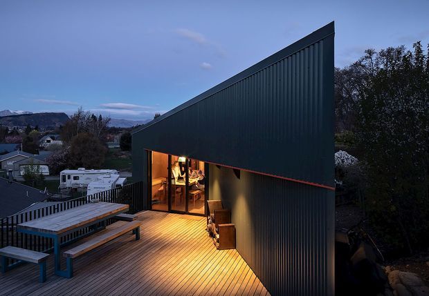 Twilight exterior view of a sharply angular, single-storey building with a steep mono-pitched roof and dark vertically ribbed metal cladding. A warm-lit glazed opening connects to a broad timber deck with built-in bench seating and outdoor furniture. The home sits in a suburban or semi-rural setting with distant hills visible beyond.